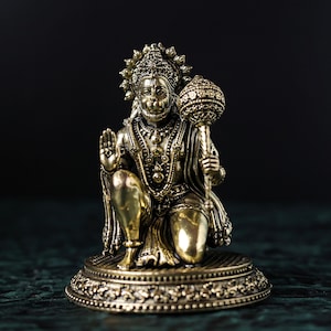 Puede incluir: Una estatua dorada del dios hindú Hanuman, sentado con la mano derecha levantada en un gesto de bendición. Sostiene una maza en su mano izquierda. La estatua está sobre una base decorativa.