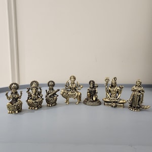 May include: A set of seven metallic gold-toned figurines depicting various deities. The collection includes figures in seated and standing poses, featuring intricate details, ornate clothing, and symbolic attributes, arranged in a horizontal row.