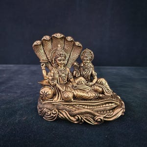 May include: A gold-colored statue of a Hindu deity couple, Vishnu and Lakshmi, sitting on a multi-headed serpent. The statue is intricately detailed and has a traditional Indian design.