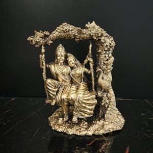 K&ouml;nnte beinhalten: Eine goldfarbene Statue eines Paares, das auf einer Schaukel sitzt, umgeben von einem Baum mit Pfauen und Blumen. Die Statue ist aus Metall und hat ein detailliertes Design.