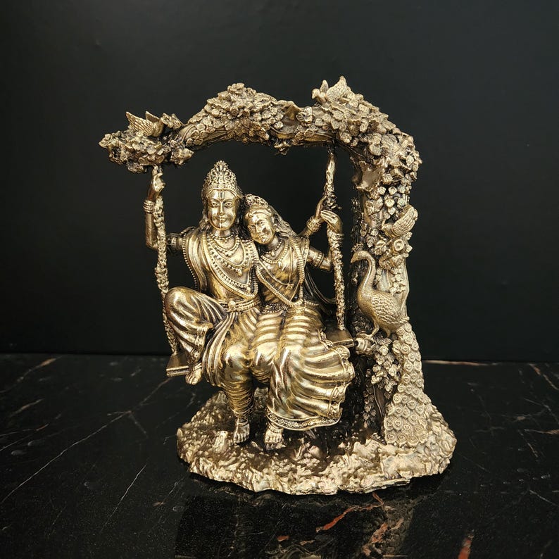 K&ouml;nnte beinhalten: Eine goldfarbene Statue eines Paares, das auf einer Schaukel sitzt, umgeben von einem Baum mit Pfauen und Blumen. Die Statue ist aus Metall und hat ein detailliertes Design.