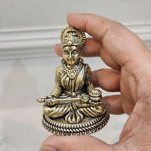 May include: A detailed brass statue of a seated deity, featuring intricate carvings and ornate details. The figure is adorned with a crown and jewelry, holding a symbolic object. The statue has a warm, golden hue and is approximately 3 inches tall.