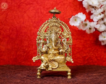 May include: A gold-colored metal statue of Ganesha, the Hindu god of wisdom and prosperity, sitting on a throne with intricate details. The statue is on a brown surface.