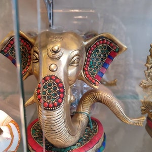 May include: A gold-colored Ganesha statue with red, green, and blue gemstone accents. The statue is in a seated position with the trunk of the elephant curled up. The statue is on a base with a multi-colored mosaic design.