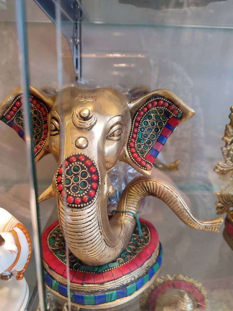 May include: A gold-colored Ganesha statue with red, green, and blue gemstone accents. The statue is in a seated position with the trunk of the elephant curled up. The statue is on a base with a multi-colored mosaic design.