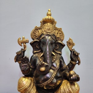 May include: A gold and black metal statue of the Hindu deity Ganesha. The statue is intricately detailed and features a large elephant head with a trunk raised in a blessing gesture. Ganesha is often depicted with four arms, holding various symbols of prosperity and good fortune.
