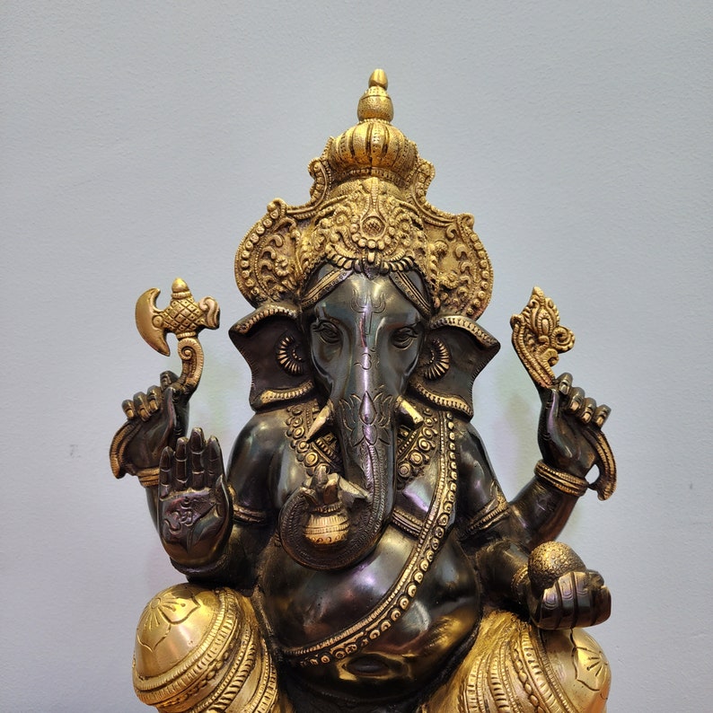May include: A gold and black metal statue of the Hindu deity Ganesha. The statue is intricately detailed and features a large elephant head with a trunk raised in a blessing gesture. Ganesha is often depicted with four arms, holding various symbols of prosperity and good fortune.