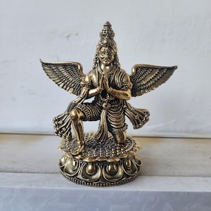 Può includere: Statua color bronzo di una figura inginocchiata con grandi ali dettagliate. La figura è adornata da una corona e gioielli, con le mani giunte in preghiera. La statua è montata su una base decorativa a motivi floreali.
