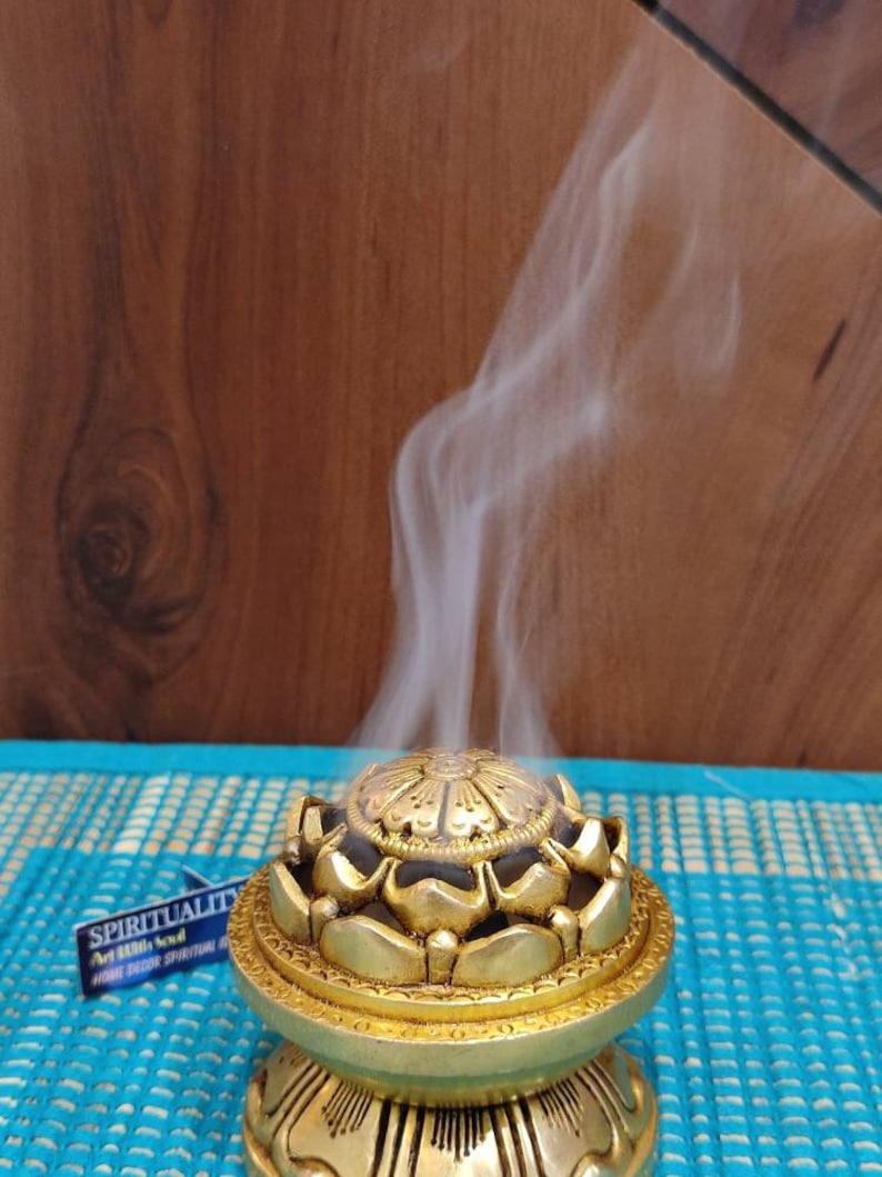 May include: A gold metal incense burner shaped like a lotus flower with smoke rising from the top. The burner is sitting on a blue and white mat.