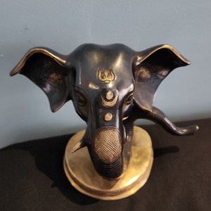 May include: A black and gold elephant head statue with intricate details. The elephant's trunk is curled up and the head is adorned with a gold symbol on the forehead.