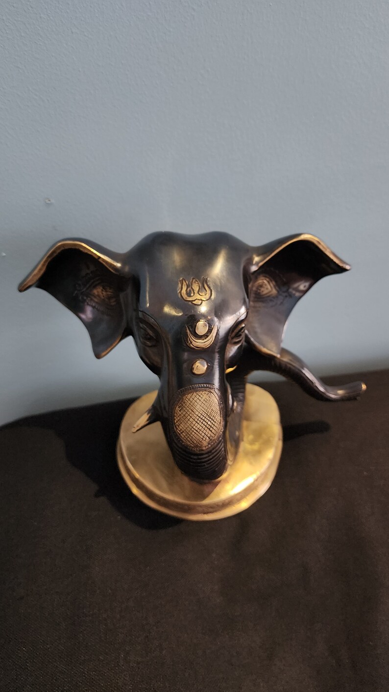 May include: A black and gold elephant head statue with intricate details. The elephant's trunk is curled up and the head is adorned with a gold symbol on the forehead.
