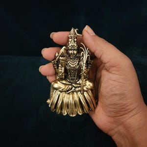 Puede incluir: Un colgante de metal dorado que representa a una deidad sentada con detalles intrincados y una corona. El colgante se sostiene en una mano sobre un fondo azul oscuro.