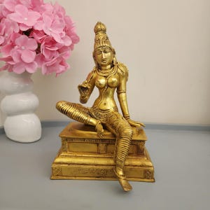 May include: A gold-toned brass statue of a seated figure with an ornate headdress, jewellery, and detailed clothing, positioned on a rectangular pedestal base with engraved patterns. The figure is depicted in a relaxed pose with one hand raised.