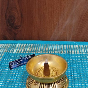 May include: A gold metal incense burner with a lotus base and a single incense stick burning in the center. The burner is on a blue and white patterned surface. The text on the label reads "SPIRITUALITYARTS Art With Soul HOME DECOR SPIRITUAL IDOLS".