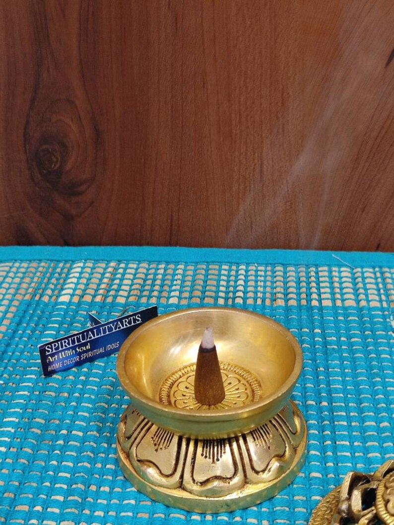 May include: A gold metal incense burner with a lotus base and a single incense stick burning in the center. The burner is on a blue and white patterned surface. The text on the label reads "SPIRITUALITYARTS Art With Soul HOME DECOR SPIRITUAL IDOLS".