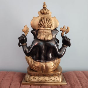 May include: A gold and black metal statue of Ganesha, the Hindu god of wisdom and prosperity. The statue is sitting on a base with intricate details.