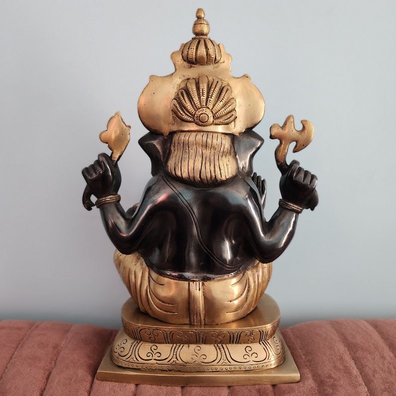 May include: A gold and black metal statue of Ganesha, the Hindu god of wisdom and prosperity. The statue is sitting on a base with intricate details.