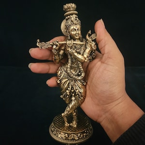 May include: A gold-colored metal statue of Krishna playing a flute. The statue is intricately detailed and features a peacock feather on the flute. The statue is standing on a round base with intricate designs.