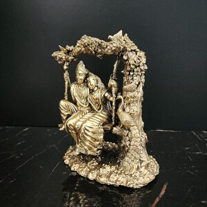 K&ouml;nnte beinhalten: Eine goldfarbene Statue eines Paares, das auf einer Schaukel sitzt, umgeben von aufwendigen floralen Details. Die Schaukel ist an einem Baumzweig aufgeh&auml;ngt, und ein Pfau sitzt auf dem Baumstamm. Die Statue ist im traditionellen indischen Stil gehalten.