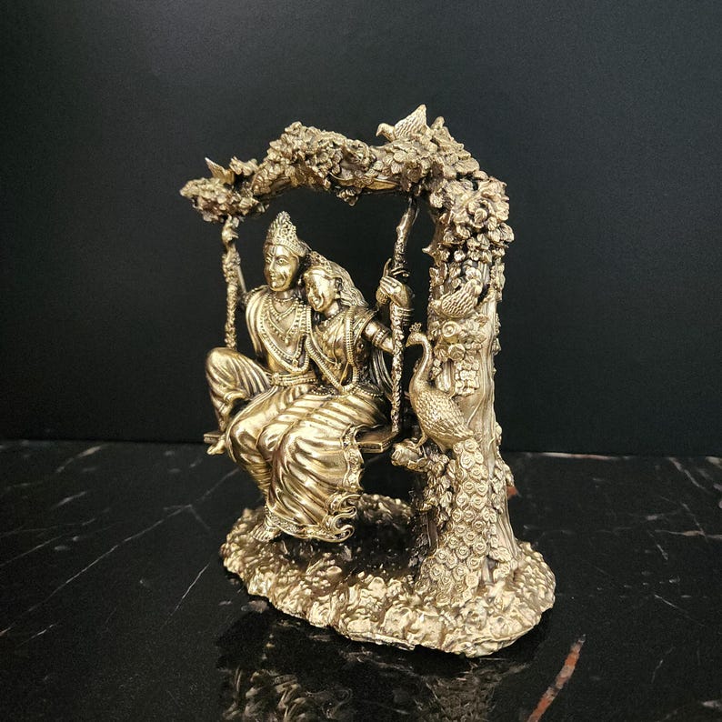 K&ouml;nnte beinhalten: Eine goldfarbene Statue eines Paares, das auf einer Schaukel sitzt, umgeben von aufwendigen floralen Details. Die Schaukel ist an einem Baumzweig aufgeh&auml;ngt, und ein Pfau sitzt auf dem Baumstamm. Die Statue ist im traditionellen indischen Stil gehalten.