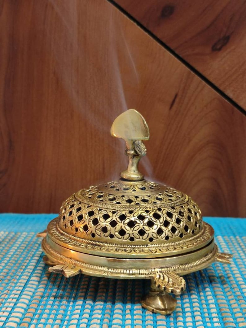 May include: A gold metal incense burner with intricate designs and a pointed top. The burner is sitting on a blue and white mat and smoke is rising from the top.