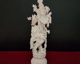 May include: White figurine of a Hindu deity playing a flute, standing on a pedestal with intricate details. The deity is adorned with floral decorations and jewelry.