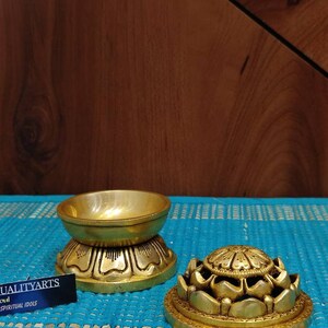 May include: Two gold-colored metal lotus flower incense burners. The top burner has a round, open top and the bottom burner has a closed top. The burners are on a blue and white patterned surface.