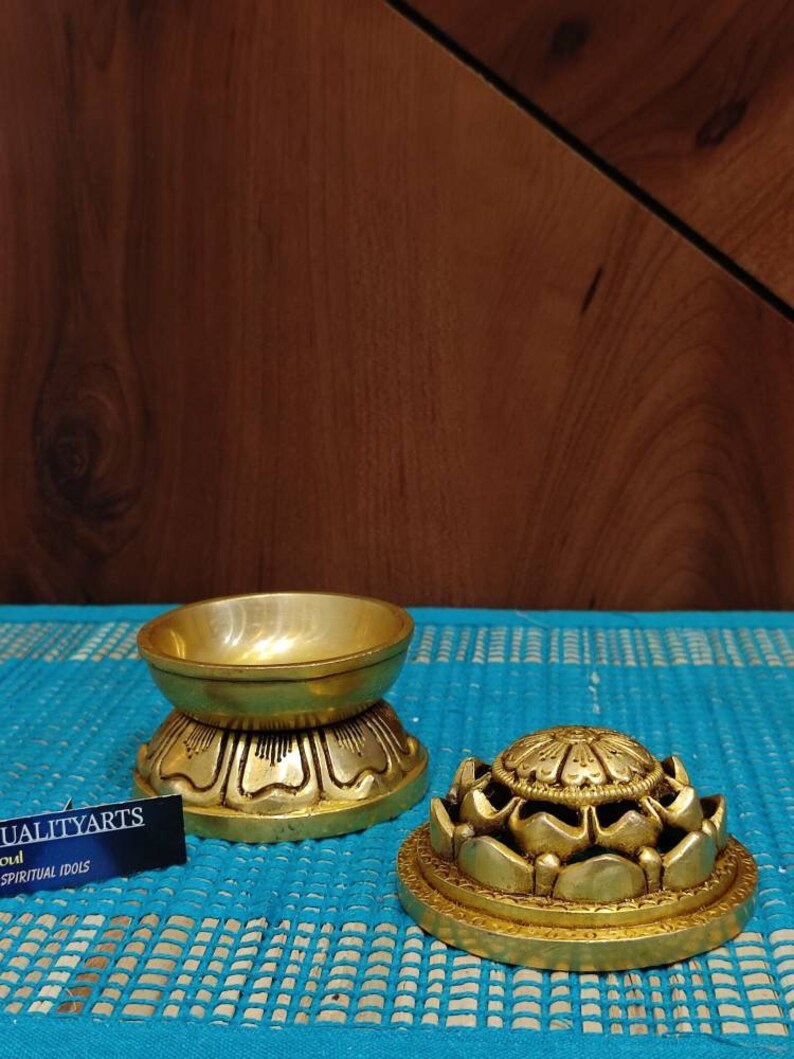May include: Two gold-colored metal lotus flower incense burners. The top burner has a round, open top and the bottom burner has a closed top. The burners are on a blue and white patterned surface.