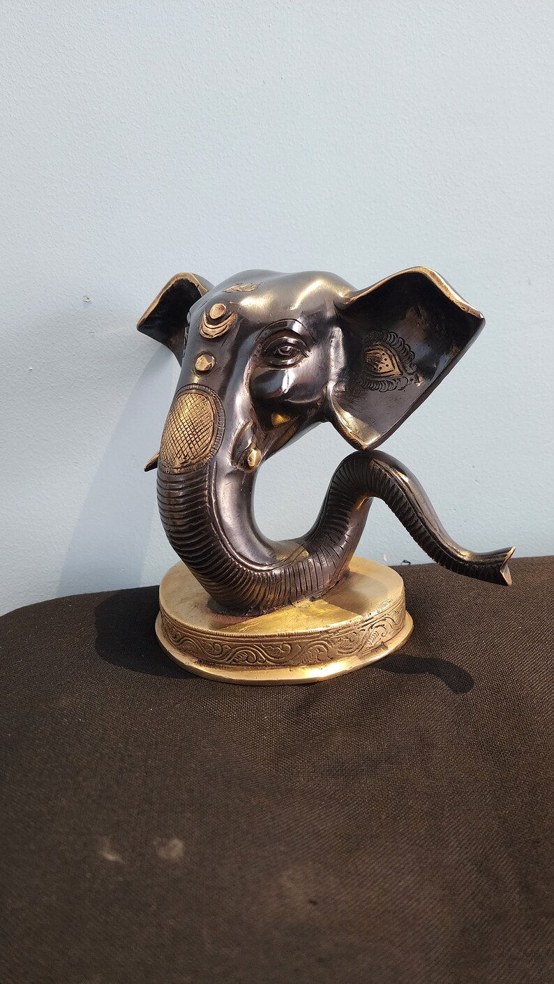 May include: A black and gold Ganesha statue with intricate details. The statue is made of metal and has a round base. The elephant's trunk is curled up and its eyes are closed.