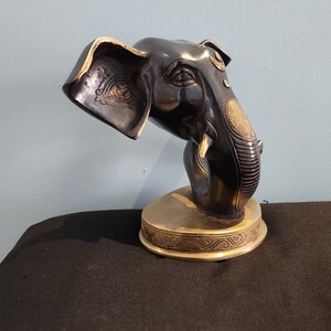 May include: A black and gold metal elephant head sculpture with intricate details. The elephant's trunk is curled up and the head is tilted to the side. The sculpture is on a gold base with a decorative design.