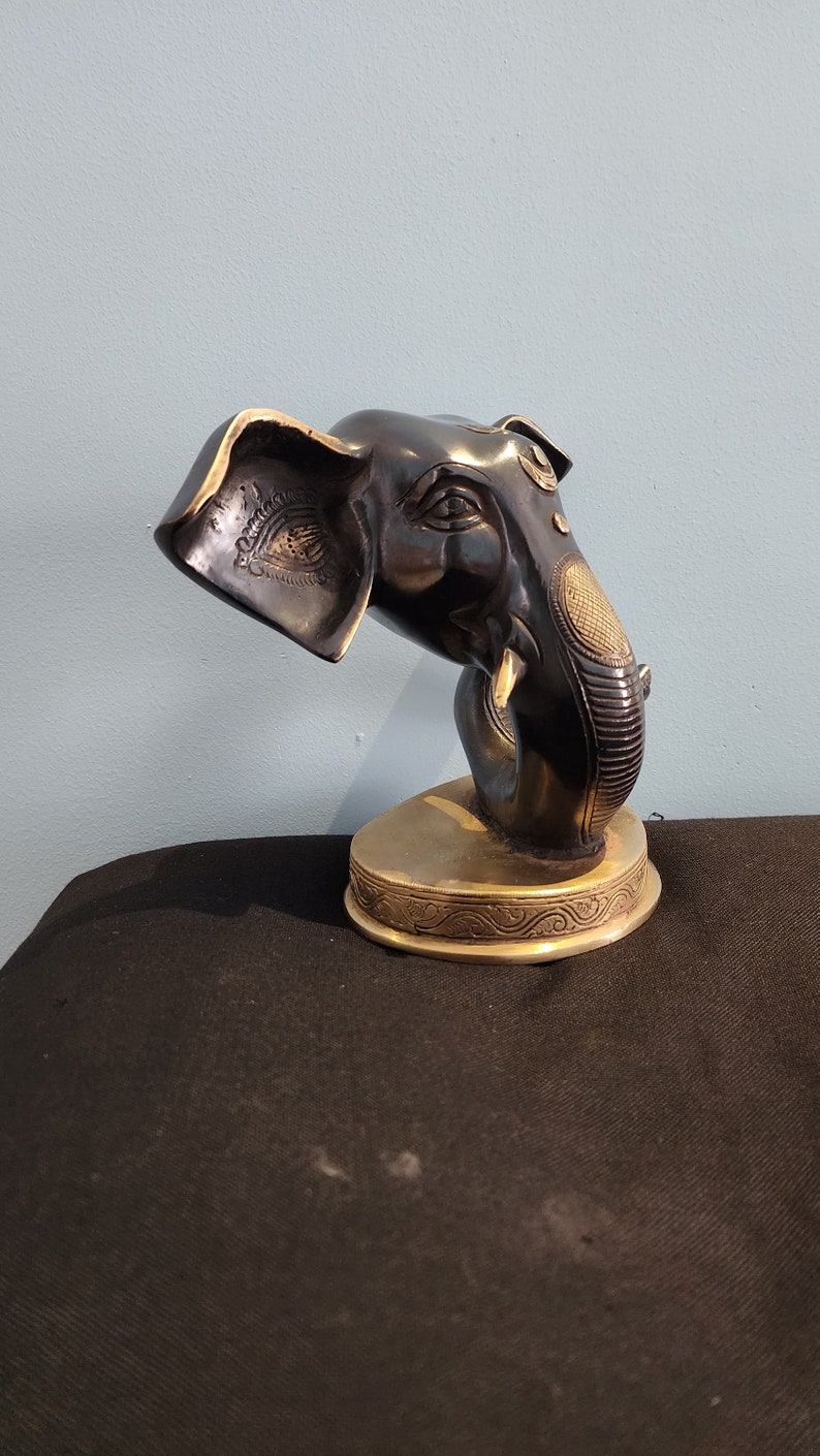 May include: A black and gold metal elephant head sculpture with intricate details. The elephant's trunk is curled up and the head is tilted to the side. The sculpture is on a gold base with a decorative design.