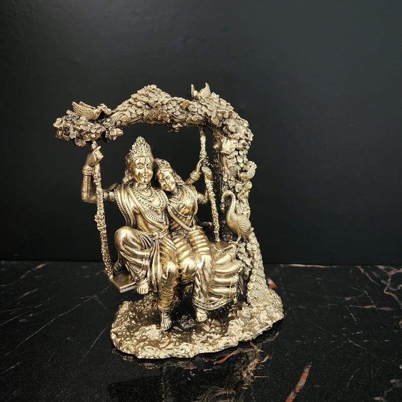 K&ouml;nnte beinhalten: Eine goldfarbene Statue eines Paares, das auf einer Schaukel sitzt, umgeben von einem Baum mit V&ouml;geln und einem Pfau. Die Statue ist aus Metall und hat ein detailliertes Design.