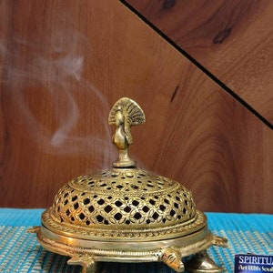 May include: A gold metal incense burner shaped like a turtle with a peacock design on top. The burner has a decorative, openwork dome and is sitting on a blue mat. Smoke is rising from the burner.