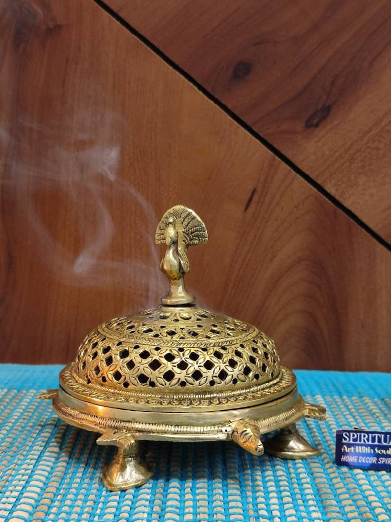 May include: A gold metal incense burner shaped like a turtle with a peacock design on top. The burner has a decorative, openwork dome and is sitting on a blue mat. Smoke is rising from the burner.
