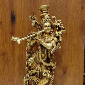 Puede incluir: Una estatua de metal dorado de Krishna tocando la flauta. La estatua está intrincadamente detallada y presenta una base decorativa.