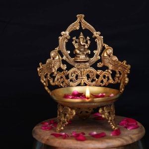 Könnte beinhalten: Eine goldene Metall-Diya mit filigranen Designs, die einen Pfau und eine Gottheit darstellen. Die Diya ist mit einer Flamme beleuchtet und von Rosenblüten umgeben.