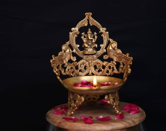 May include: A gold metal diya with intricate designs featuring a peacock and a deity figure. The diya is lit with a flame and surrounded by rose petals.