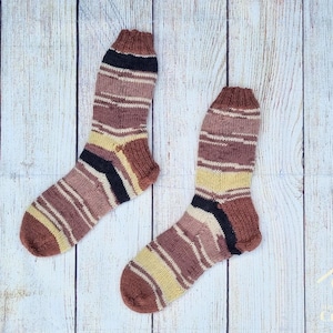 May include: A pair of hand-knitted socks featuring a variegated striped pattern in shades of brown, tan, cream, and black. The socks have a ribbed cuff at the top and a reinforced heel and toe section, showcasing a consistent knit texture throughout.