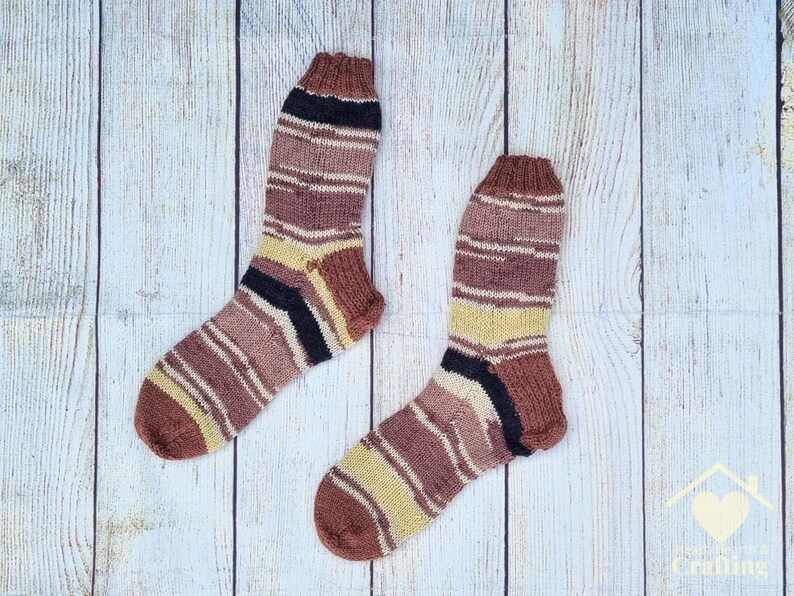 May include: A pair of hand-knitted socks featuring a variegated striped pattern in shades of brown, tan, cream, and black. The socks have a ribbed cuff at the top and a reinforced heel and toe section, showcasing a consistent knit texture throughout.