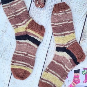 May include: A pair of hand-knitted socks featuring horizontal stripes in shades of brown, cream, yellow, and black. The socks have a ribbed cuff and a reinforced heel construction. A small portion of a craft instruction card is visible.