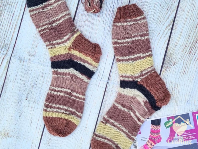 May include: A pair of hand-knitted socks featuring horizontal stripes in shades of brown, cream, yellow, and black. The socks have a ribbed cuff and a reinforced heel construction. A small portion of a craft instruction card is visible.