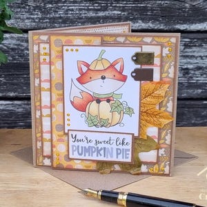 Peut inclure: Une carte de v&oelig;ux artisanale illustr&eacute;e d'un renard portant un chapeau citrouille et assis sur une citrouille. La carte pr&eacute;sente du papier &agrave; motifs automnaux, une grande feuille d'&eacute;rable en tissu et le texte "You're sweet like PUMPKIN PIE".
