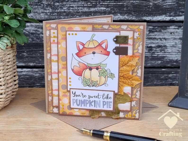 Peut inclure: Une carte de v&oelig;ux artisanale illustr&eacute;e d'un renard portant un chapeau citrouille et assis sur une citrouille. La carte pr&eacute;sente du papier &agrave; motifs automnaux, une grande feuille d'&eacute;rable en tissu et le texte "You're sweet like PUMPKIN PIE".