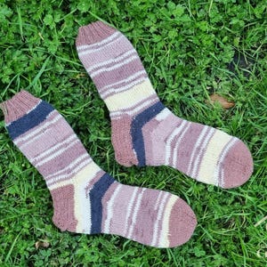 May include: A pair of hand-knitted socks featuring horizontal stripes in shades of mauve, cream, and dark navy blue. The socks have a ribbed cuff, a reinforced heel, and a rounded toe. A logo with the text Heart, Hearth & Crafting is visible.