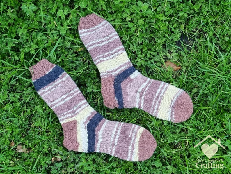 May include: A pair of hand-knitted socks featuring horizontal stripes in shades of mauve, cream, and dark navy blue. The socks have a ribbed cuff, a reinforced heel, and a rounded toe. A logo with the text Heart, Hearth & Crafting is visible.