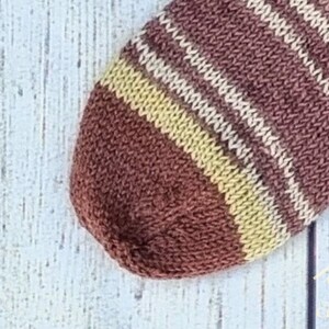 May include: A hand-knitted sock featuring a rounded toe section. The fabric displays a repeating pattern of horizontal stripes in shades of deep brown, muted tan, and cream, created with a consistent stockinette stitch texture.