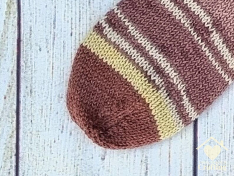 May include: A hand-knitted sock featuring a rounded toe section. The fabric displays a repeating pattern of horizontal stripes in shades of deep brown, muted tan, and cream, created with a consistent stockinette stitch texture.