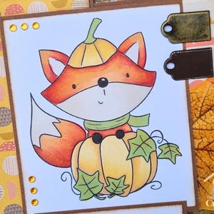 Peut inclure: Une carte de v&oelig;ux artisanale avec une illustration color&eacute;e d'un renard portant un chapeau citrouille et assis sur une citrouille. La carte comprend des feuilles vertes, des ornements dor&eacute;s et deux &eacute;tiquettes m&eacute;talliques bronze et brun.