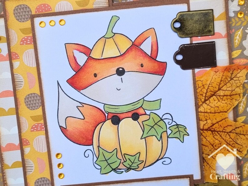 Peut inclure: Une carte de v&oelig;ux artisanale avec une illustration color&eacute;e d'un renard portant un chapeau citrouille et assis sur une citrouille. La carte comprend des feuilles vertes, des ornements dor&eacute;s et deux &eacute;tiquettes m&eacute;talliques bronze et brun.