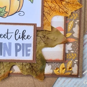Peut inclure: Une carte de v&oelig;ux artisanale avec un design superpos&eacute;, une illustration de citrouille et le texte "sweet like PUMPKIN PIE". La carte est d&eacute;cor&eacute;e de feuilles d'automne en tissu dans des tons orange et marron, de papier &agrave; motifs et de trois petites gemmes adh&eacute;sives ambr&eacute;es.